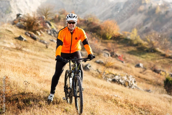 Fototapeta Man in helmet and glasses stay on the bicycle under landscape with rocks and hill.
