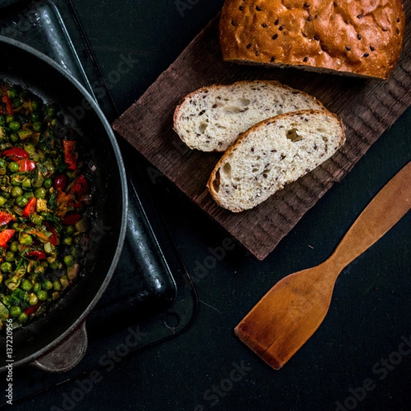 Fototapeta Vegetable stew with green peas, red pepper, onion and zucchini and spices. In a frying pan on a black background. On a wooden platform grain bread with flax seeds. Size 1 to 1.