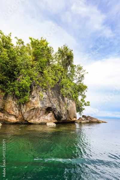Obraz Tropical rock island covered with palm trees and dense jungle rises from the calm waters of Triton Bay, West Papua, Indonesia. A stunning seascape surrounded by untouched rainforest and cloudy skies.