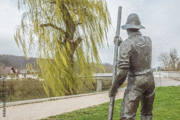 Obraz Sturmbewegte Trauerweide neben Skulptur eines Flößers vor Fußgängersteg in Neckartenzlingen