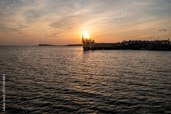 Fototapeta Silhouetted figures stand against setting sun at Blackrock platform as golden light ripples on water.