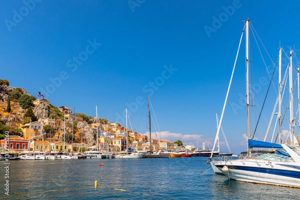 Fototapeta Simi Island harbor view with sailboats anchored, hillside town with colorful buildings.  Blue sky above, calm water reflecting the scene.
