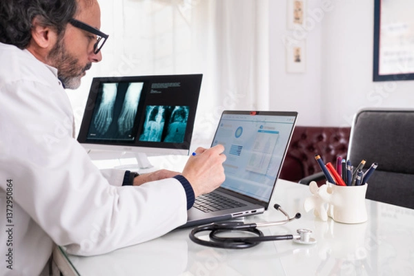 Fototapeta A middle-aged doctor wearing a white coat studies a case using generic anonymous healthcare software. In the background, there is another screen with some X-rays and a corridor