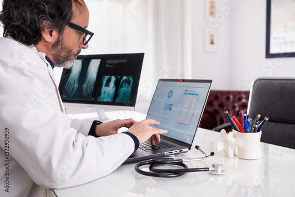 Fototapeta A middle-aged doctor wearing a white coat types on his laptop using generic anonymous healthcare software. In the background, there is another screeen with some X-rays and a corridor