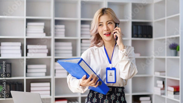 Fototapeta Professional businesswoman talking on smartphone while holding project folders in modern office
