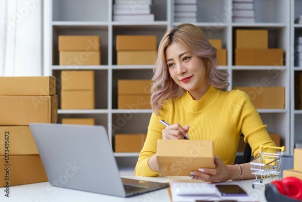 Fototapeta Cheerful female online seller talking on phone while preparing delivery boxes in home office workspace