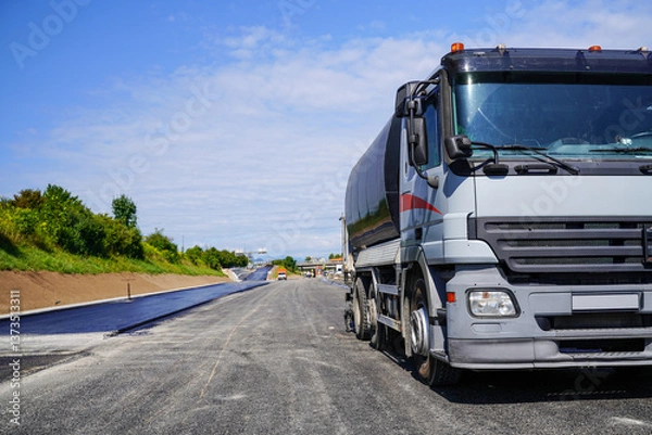 Fototapeta Truck with bitumen spreader attachment applying hot bitumen coating on fresh asphalt with highway construction in the background