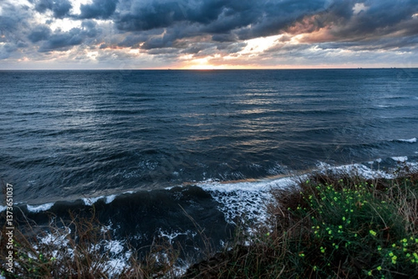 Obraz The ocean is calm and the sky is cloudy. The sun is setting and the water is reflecting the colors of the sky