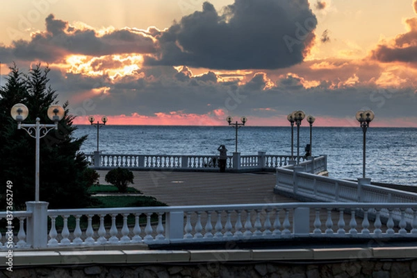Obraz A beautiful sunset over the ocean with a pier in the background. The sky is filled with clouds and the water is calm. The pier is lit up with lights, creating a serene atmosphere