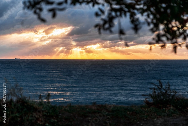 Obraz Gelendzhik resort. The sun is setting over the ocean, casting a warm glow on the water. The sky is filled with clouds, creating a moody atmosphere