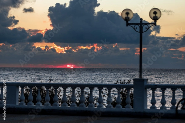 Obraz The sky is filled with clouds and the sun is setting. The water is calm and the sky is a beautiful orange color