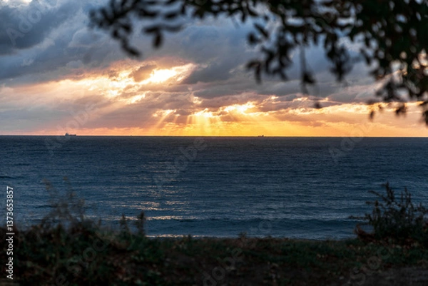 Obraz Gelendzhik resort. The sun is setting over the ocean, casting a warm glow on the water. The sky is filled with clouds, creating a moody atmosphere