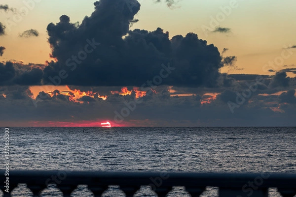 Obraz The sky is filled with clouds and the sun is setting. The water is calm and the sky is a beautiful orange color