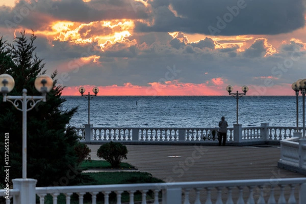 Obraz A beautiful sunset over the ocean with a pier in the background. The sky is filled with clouds and the water is calm. The pier is lit up with lights, creating a serene atmosphere