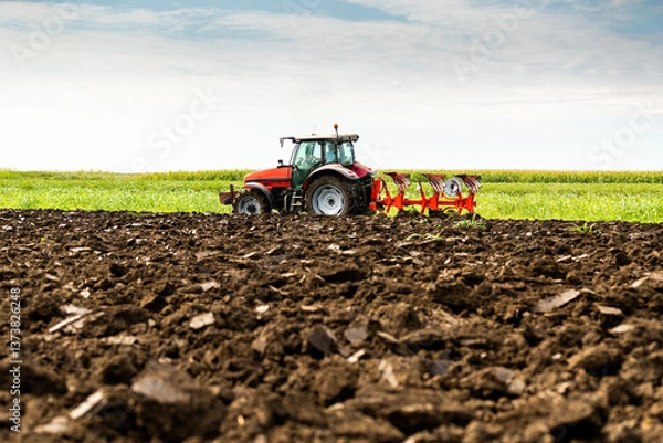 Obraz Tractor with plow attachment turns the soil in a vast, green farmland