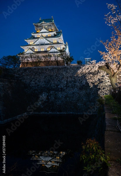 Fototapeta Osaka Castle glows under the twilight sky. Surrounding cherry blossoms illuminate the scene, creating a breathtaking spectacle of Japanese history and natures beauty.