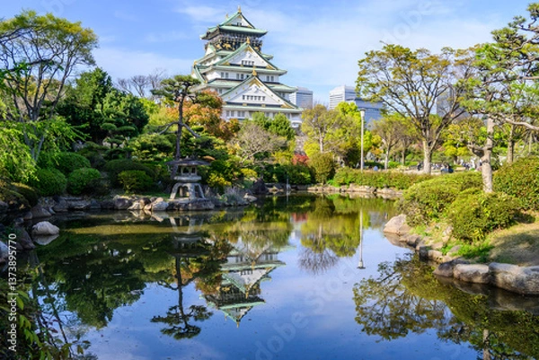 Fototapeta Osaka Castle, rebuilt in 16th century, reflects in a calm pond. Surrounding Japanese garden showcases meticulously landscaped trees and stonework. A tranquil scene.