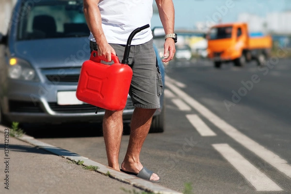 Fototapeta A man is standing by the roadside while holding a bright red gas can, looking for help