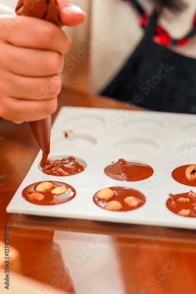 Obraz A person uses a piping bag to fill chocolate molds, with some containing nuts. This process showcases homemade chocolate making, perfect for culinary enthusiasts