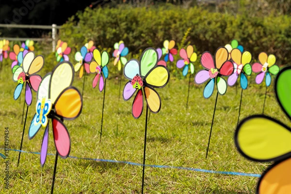Obraz A lively field of colorful pinwheels spins on green grass, enhanced by butterfly and ladybug decorations. Bright hues and a playful vibe are framed by bushes and a charming fence