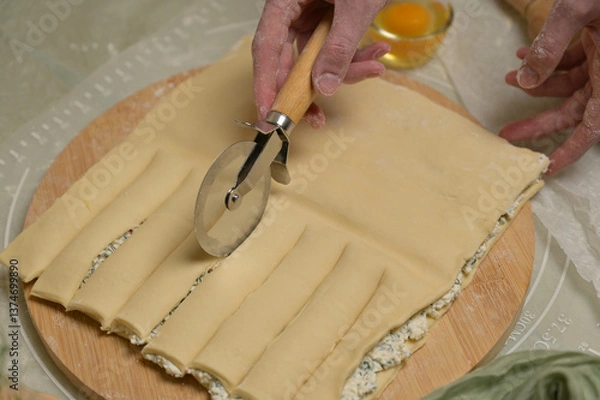 Obraz Close up square sheet of dough cut into strips with woman's hands on wooden board. Cutter, egg on kitchen table. Home cooking baking puffs