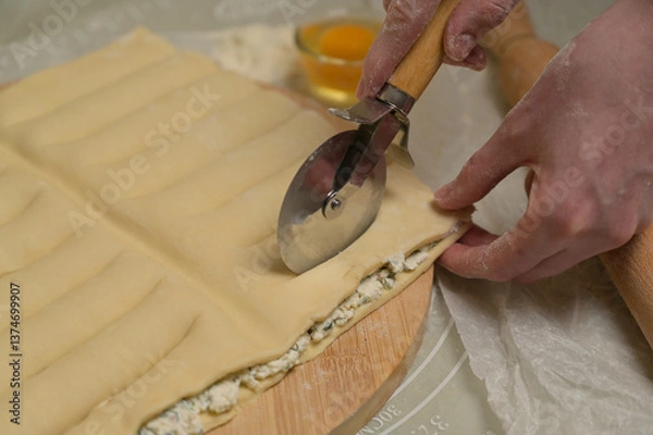 Obraz Close up square sheet of dough cut into strips with woman's hands on wooden board. Cutter, flour, egg on kitchen table. Home cooking baking puffs