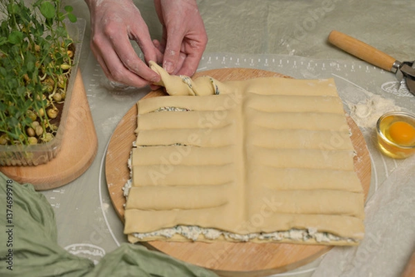 Obraz Woman's hands twists dough strips on wooden board. Cutter, egg, green towel, microgreens sprouts on kitchen table. Home cooking baking puffs