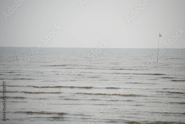 Fototapeta An expansive, tranquil seascape unfolds under a soft, overcast sky, featuring gently rippling waters punctuated by sparse, distant markers and a lone flag. This serene scene conveys a sense of calm