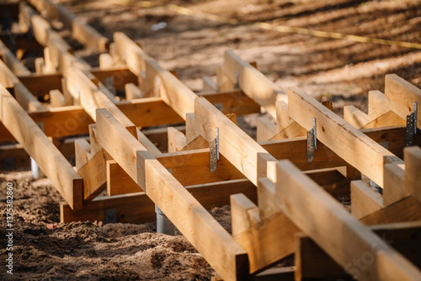 Obraz Close-up of wooden staircase framework under construction on sandy ground, showing beams, brackets, and metal supports in a forested outdoor setting..