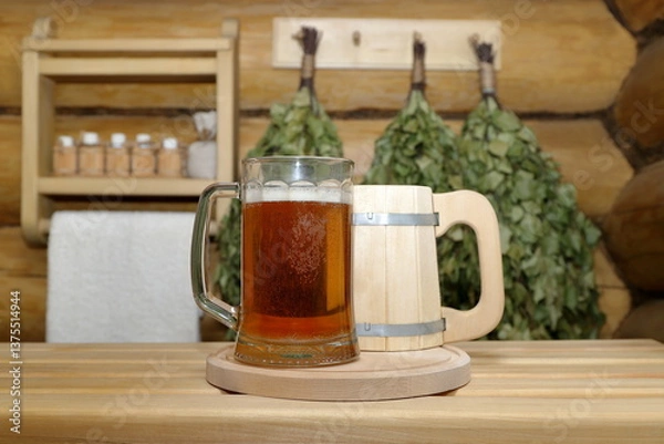 Fototapeta In the sauna interior, on a plank bench a glass mug of light beer stands next to a wooden mug on the background of a log wall with dry birch brooms and shelf with bottles of aromatic oils and towel.