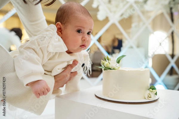 Fototapeta A minimalist white wedding cake with a smooth buttercream finish, adorned with delicate white flowers and greenery, displayed on a table.