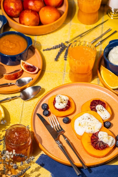 Fototapeta Sliced persimmon with ricotta and blood orange on colorful table setting