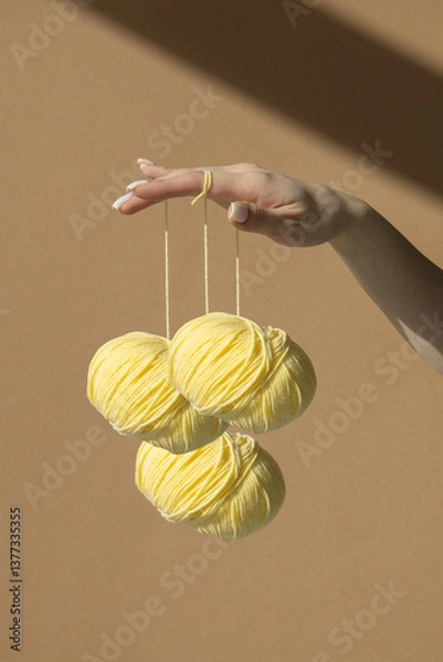 Obraz Three yellow balls of knitting thread hang on a woman's beautifully manicured fingers. Wool skeins with yarn on a light brown background in sunlight.