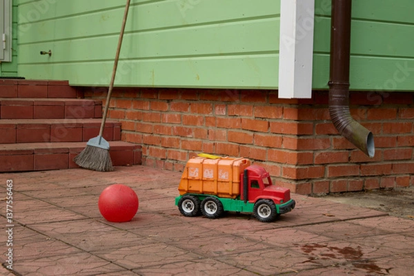 Obraz Children's toy truck and red ball placed on a brick patio near a broom and green siding. Scene depicts an everyday outdoor playtime setting with simple toys.