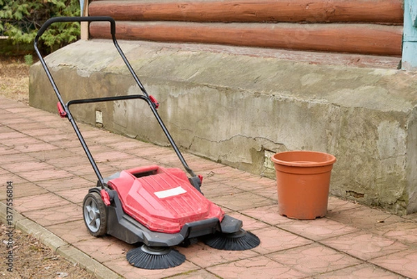 Obraz Red outdoor power sweeper rests on a paved surface with black bristles visible, accompanied by an orange bucket, set against a wooden and stone building exterior.