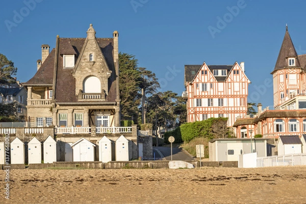 Obraz A beach at low tide with wet sand that reflects the buildings with traditional architecture  A row of beach huts typical of seaside resorts at Saint-Cast-Le-Guildo in Brittany, France