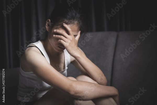Fototapeta A depressed young woman sits sadly on a sofa, covering her face with her hands. Depressing atmosphere. Relationship problems. Violence against women.