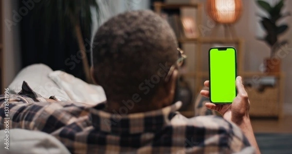 Fototapeta African American man lying at sofa and holding chroma key phone. Man having video chat lying at couch. Male relaxing at comfortable home. Man holding smartphone with green template gadget.