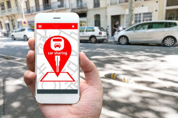Fototapeta Car sharing service or rental concept. Sharing economy and collaborative consumption. Customer hand holding smart phone with icons application screen and blur car background.