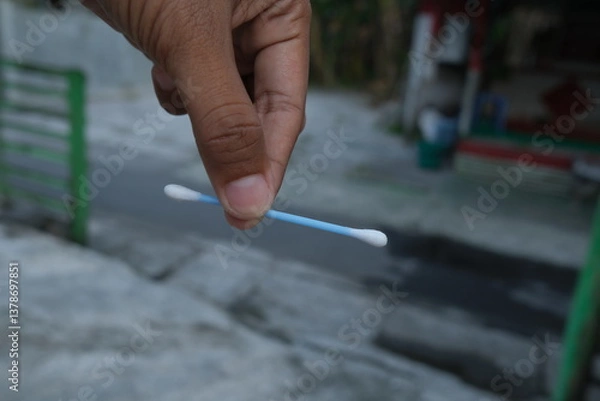 Fototapeta A hand holds a cotton swab.  The swab is light blue and white.  Outdoor setting with blurred background
