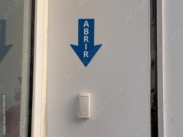 Fototapeta A blue sign with the word abrir and a downward arrow is mounted on a white wall beside a doorbell. This simple design clearly communicates the action to open the door