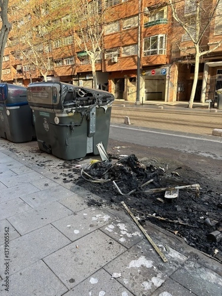 Fototapeta A city street shows a damaged trash bin, charred and smoldering, with burnt debris scattered on the sidewalk, indicating recent fire activity. Nearby buildings provide urban context