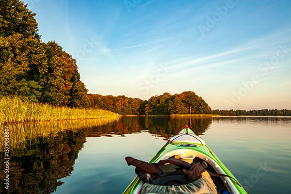 Fototapeta Kayak gliding across a tranquil lake, trees and reeds bathed in the golden light of the setting sun. 