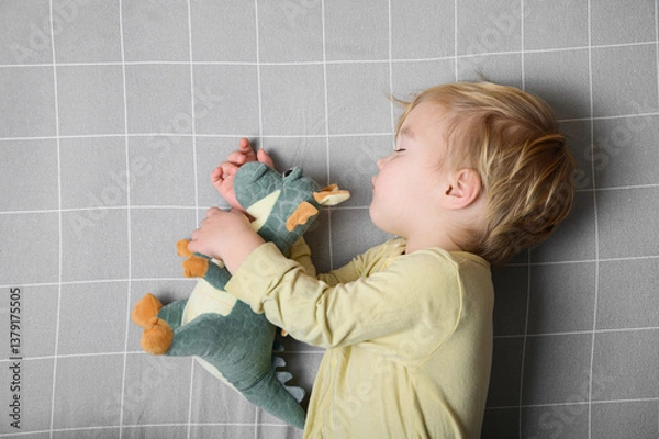 Fototapeta Sleeping Toddler Hugging Plush Dinosaur on Grey Grid Bed
