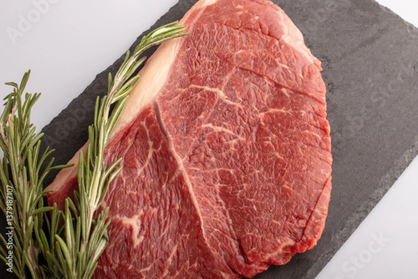 Fototapeta A thick ribeye steak with rich marbling is placed on a dark slate board alongside fresh rosemary sprigs, ideal for grilling or roasting. Perfect for a delicious meal