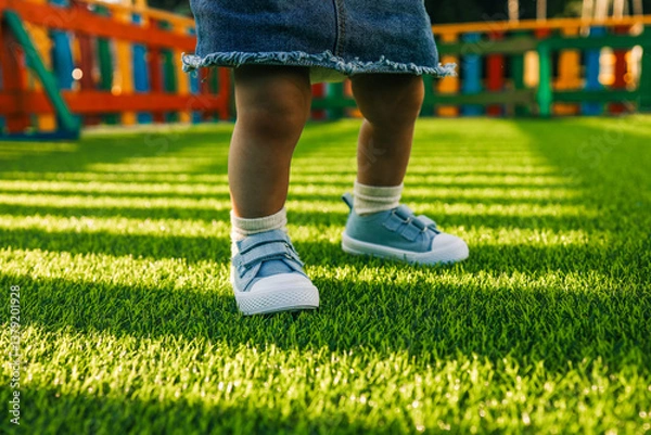 Obraz Toddler in blue sneakers standing on bright green artificial grass in a colorful playground. Low angle view of children's legs on a sunny day