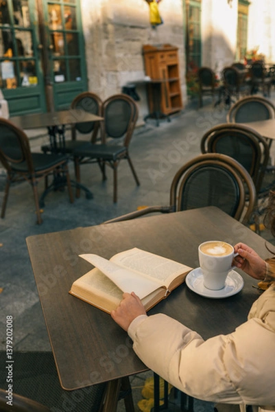 Obraz A person enjoys a latte and a book at an outdoor café table on a quiet street. Cozy autumn or winter vibe with warm light and European-style architecture in the background