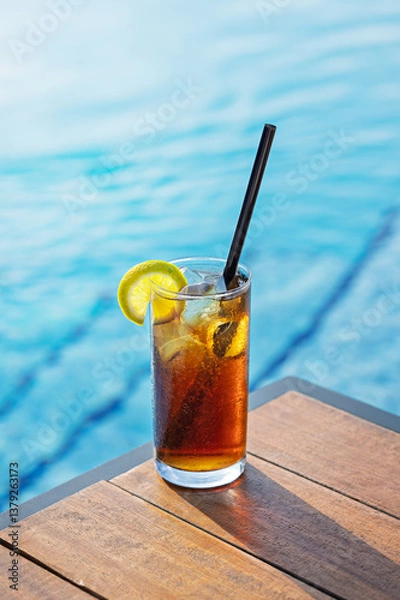 Fototapeta Cuba Libre cocktail with ice, a lemon slice, and a straw on a wooden table near a blue pool.