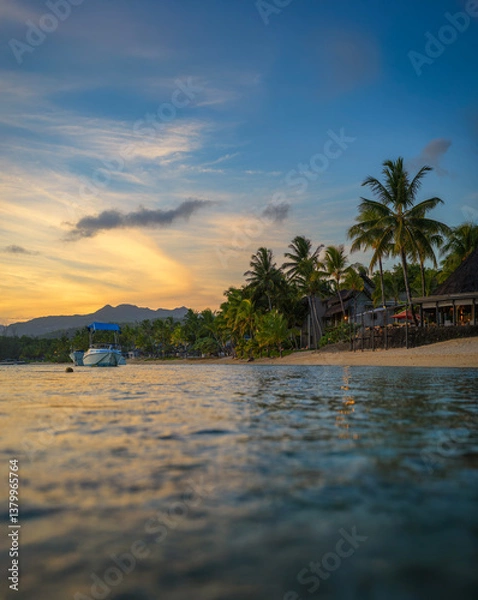 Fototapeta Serene beach in Bel Ombre, Mauritius, at sunset with calm waters, a boat floating near the shore, palm trees, beachfront resort huts, and distant mountains under a colorful sky.