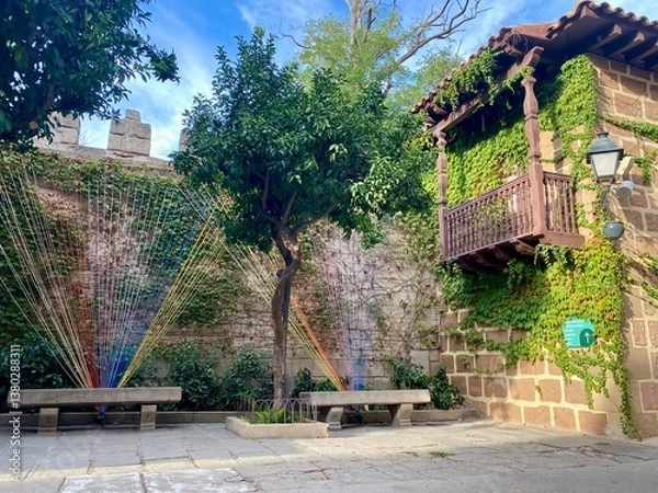 Fototapeta A peaceful courtyard featuring a rustic ivy-covered house, stone benches, lush trees, colorful yarn art installation, and playful recycling bins.